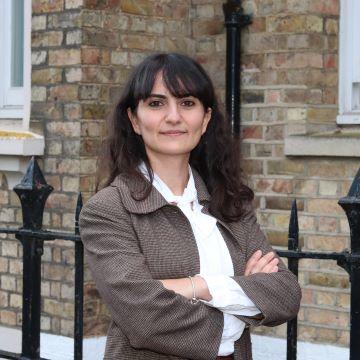 Merve square Merve is smiling and standing with her arms folded in front of a brown brick building. She has long dark hair and a fringe. She is wearing a white shirt and brown blazer.